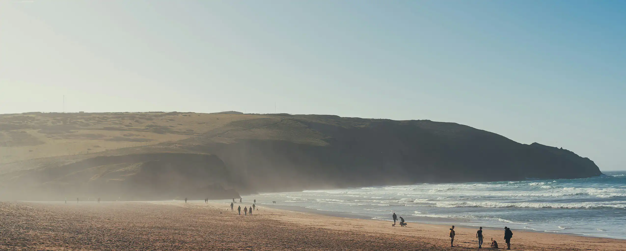 People stroll along a sandy beach in Cornwall, with the ocean waves gently lapping at the shore, a great part of the Cornish outdoors.