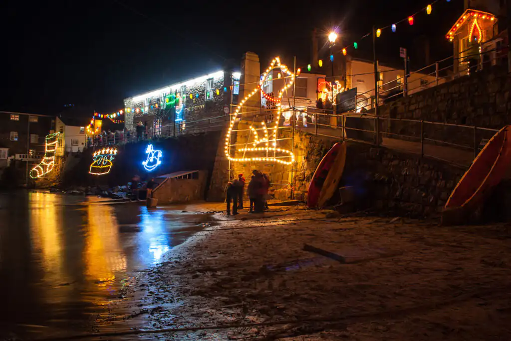People walking on a beach at night, illuminated by Mousehole Christmas lights, celebrating the New Year in Cornwall.