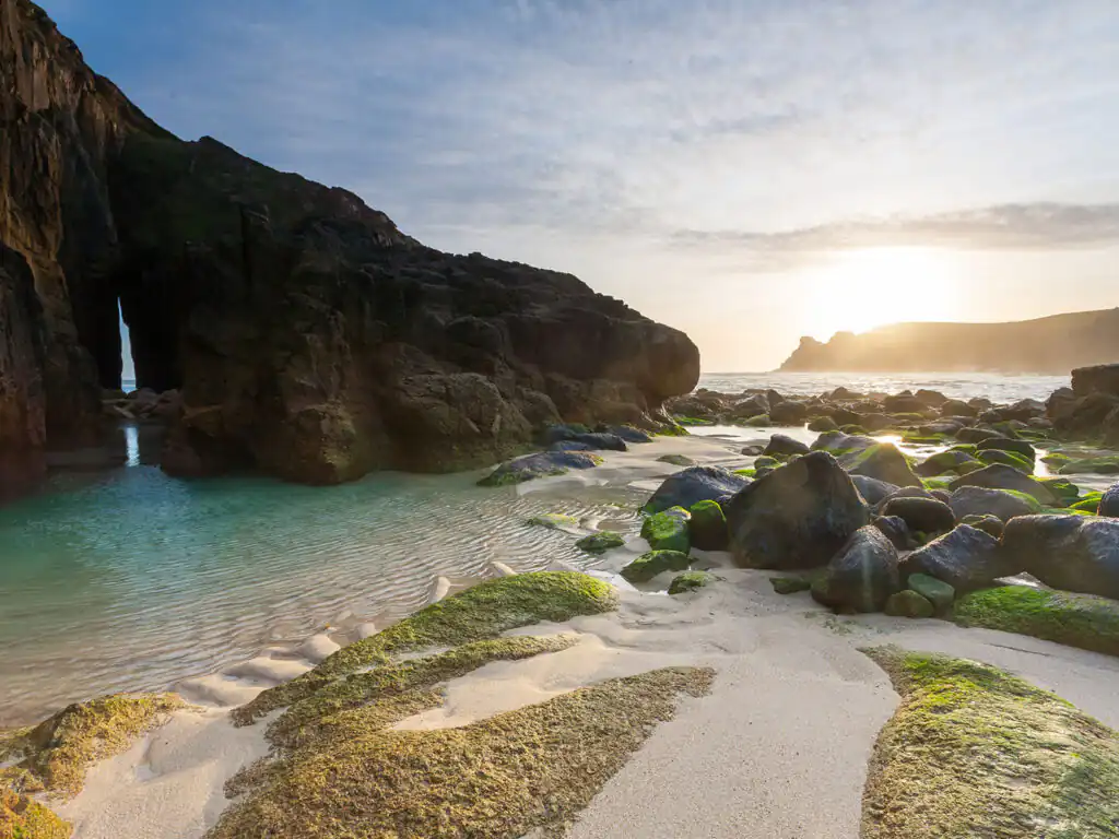 A scenic view of Nanjizal Beach featuring rocks and green algae on the sandy shore, a top beach in Cornwall.