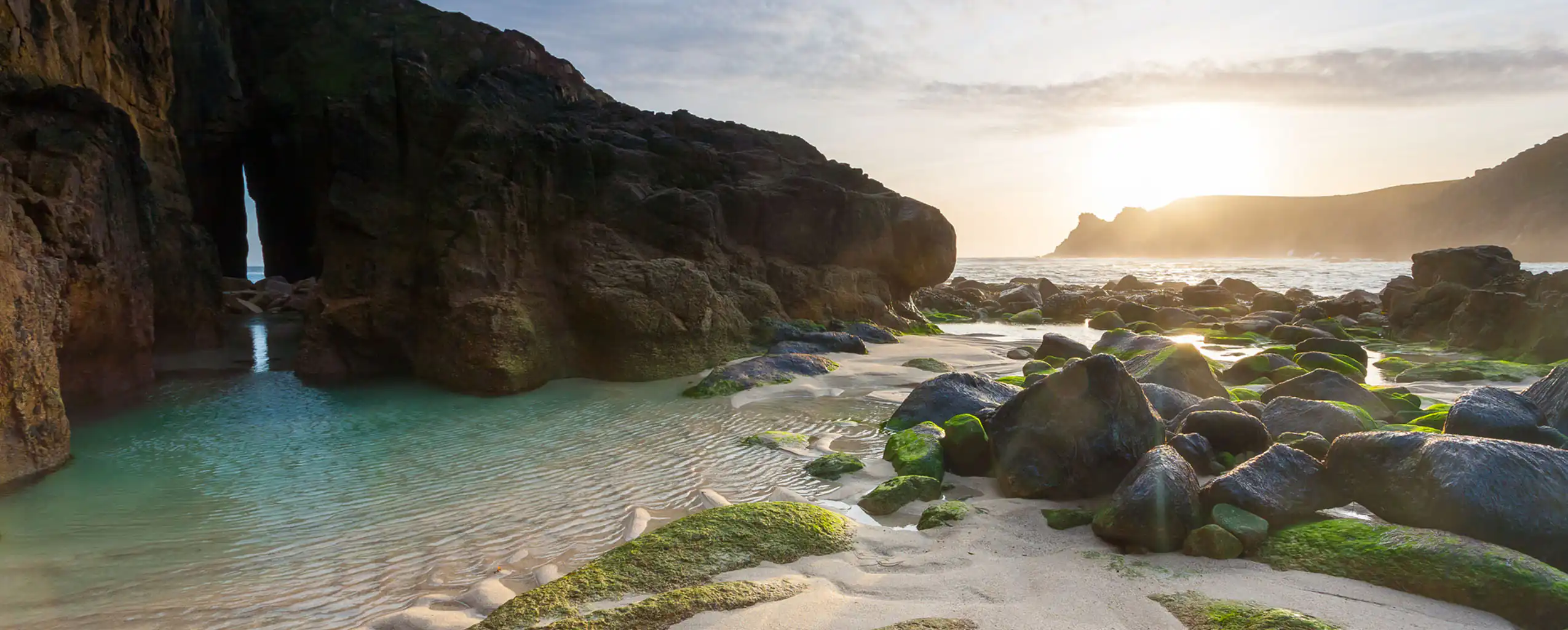 A scenic view of Nanjizal Beach featuring rocks and green algae on the sandy shore, a top beach in Cornwall.