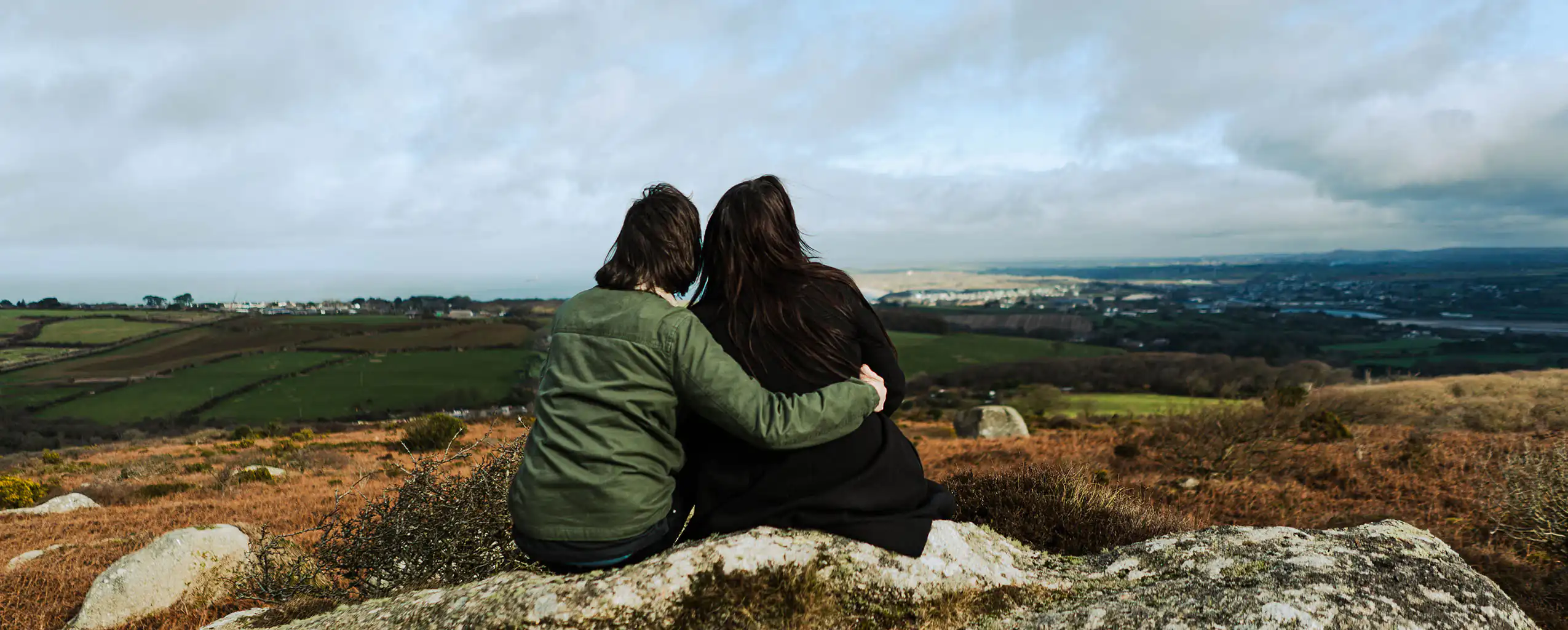 A couple sits on Trencrom Hill, gazing at the ocean, enjoying a scenic view during their holiday at a February half term cottage in Cornwall.