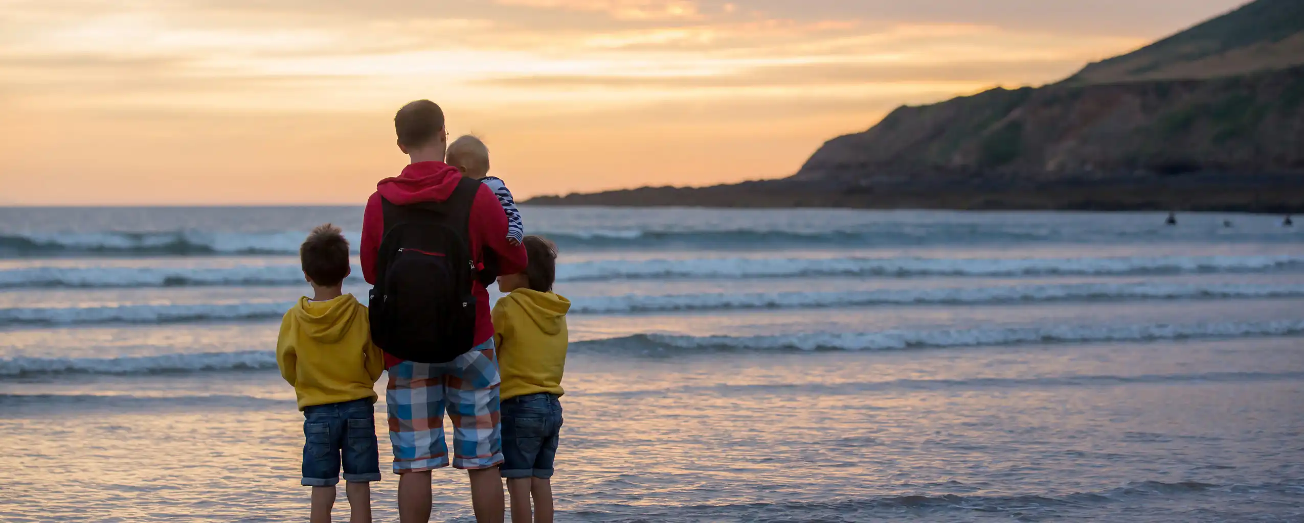 A man and two children enjoy a sunset on the beach, whilst on holiday at their family-friendly cottage in Cornwall.