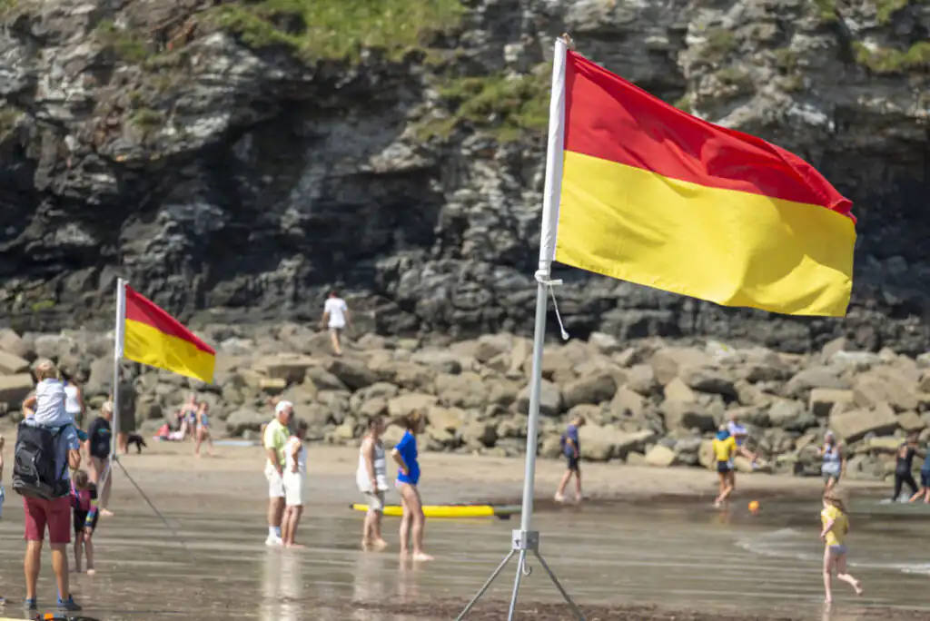 A group of beachgoers enjoying the sun at St Agnes Beach, with RNLI flags and a lifeguard on duty nearby.