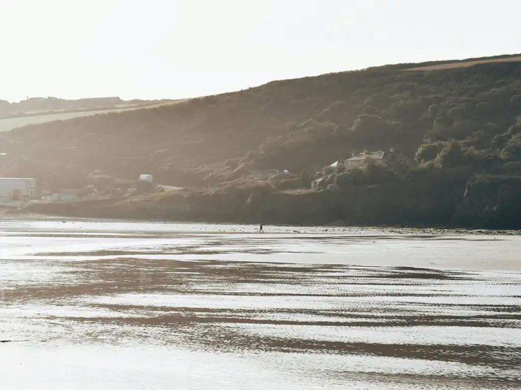A person walks along Mawgan Porth Beach, with waves gently crashing in the background, a place to visit from a Mawgan Porth Holiday Cottage.