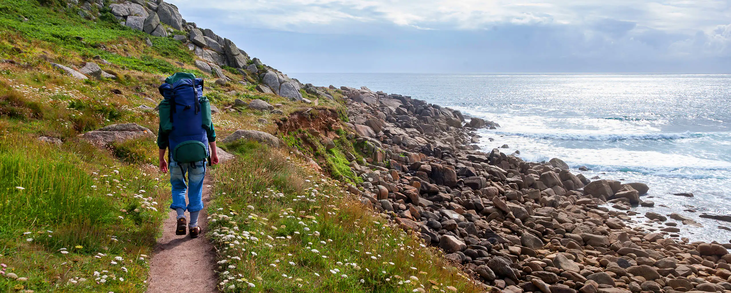 A woman walks along the South West Coastal Path, with the ocean visible beside her in beautiful Cornwall.