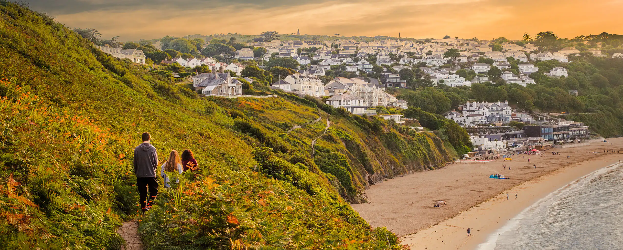Three people walk on a coastal path, overlooking the scenic Carbis Bay beach.