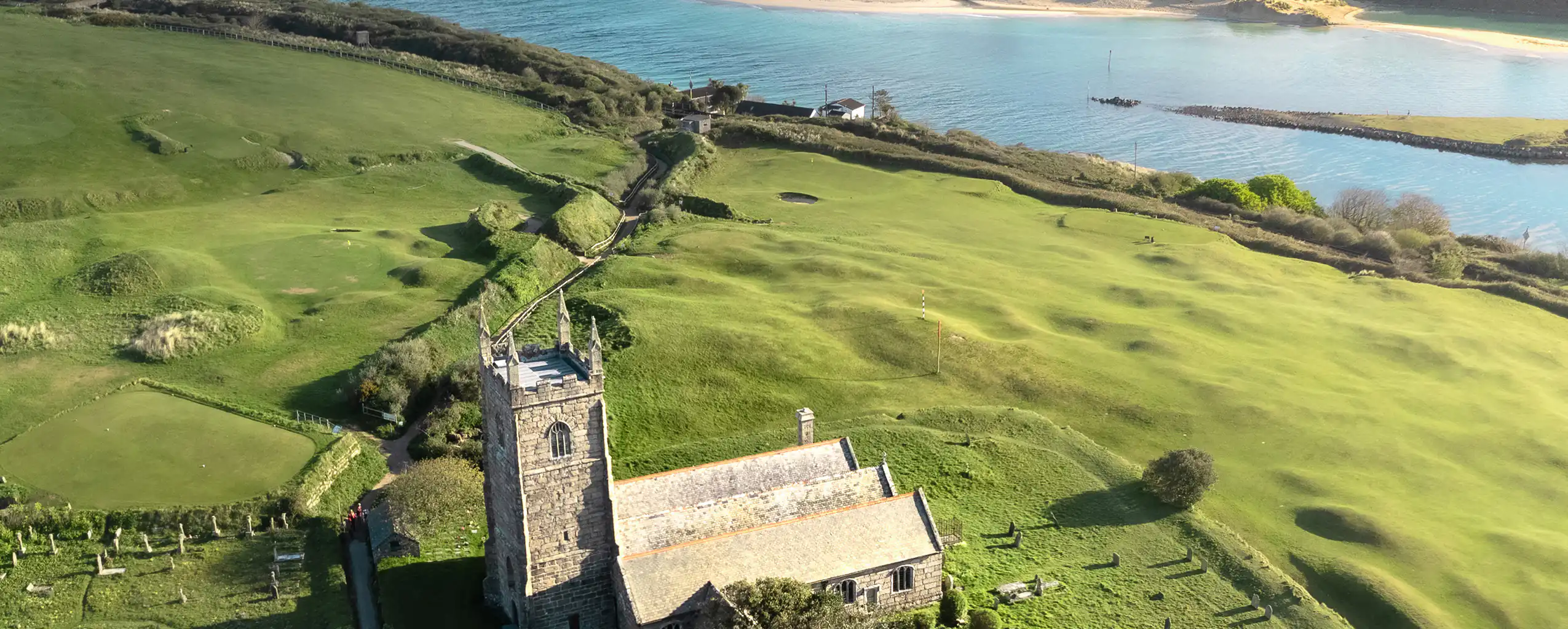Aerial view of St Uny Church on a hill, overlooking the ocean in Lelant, surrounded by lush greenery and blue waters, a great place to visit from a Lelant Holiday Cottage.