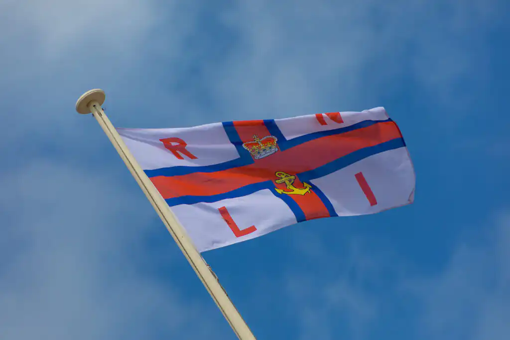 A vibrant blue sky with white clouds, showcasing the RNLI flag fluttering in the breeze in Cornwall.