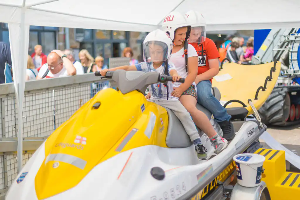 Three children enjoy a jet ski ride on a yellow and white jet ski during an RNLI demonstration in Cornwall.