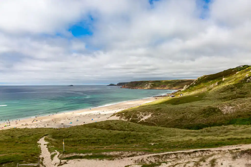 Sennen Cove beach with lush grassy hills and sparkling blue water, showcasing one of Cornwall's best surf spots.