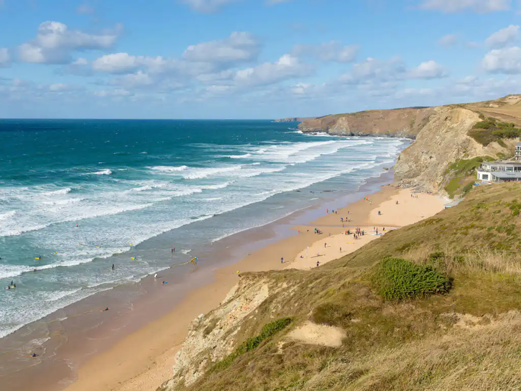 A sunny beach at Watergate Bay, Cornwall, with a few people relaxing and surfers riding the waves in the background.