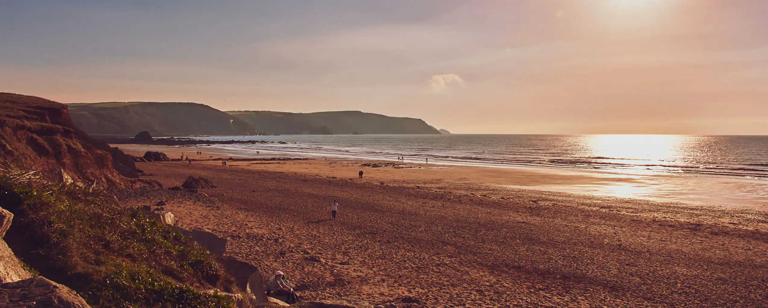 The sun sets over Widemouth Beach in Cornwall, casting warm hues across the sandy shore and tranquil waves, great for a walk from a Cornwall cottage sleeping 6.