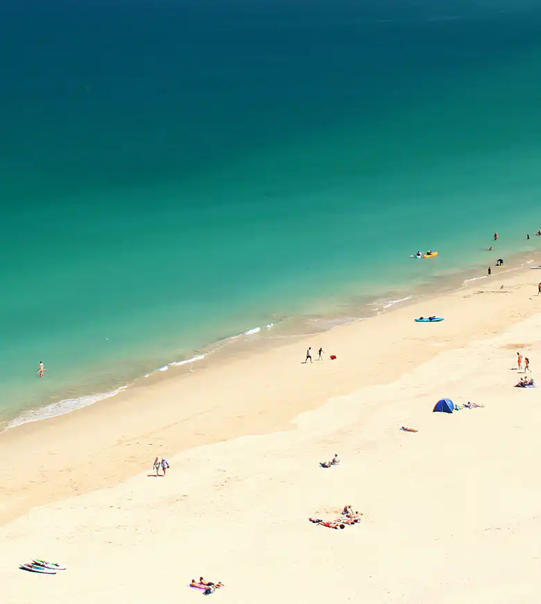 Carbis Bay Beach with sunbathers enjoying the sand and a calm body of water in the background on a sunny day.