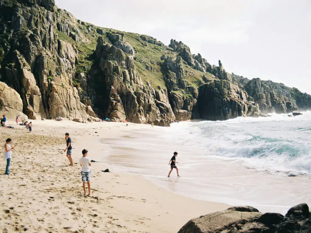 A familyrelaxing on Pednvounder Beach in Cornwall, with a dramatic rocky cliff nearby, a great place to visit during a family holiday in Cornwall.