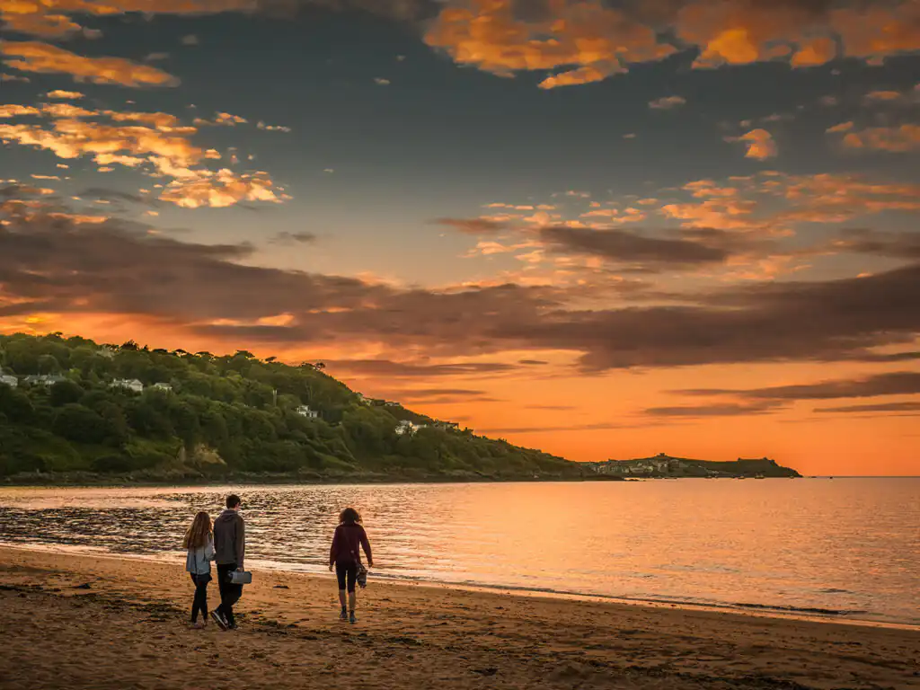 Three people stroll along Carbis Bay Beach, silhouetted against a vibrant sunset sky, minutes away from the Cornish Escapes Office in Carbis Bay.