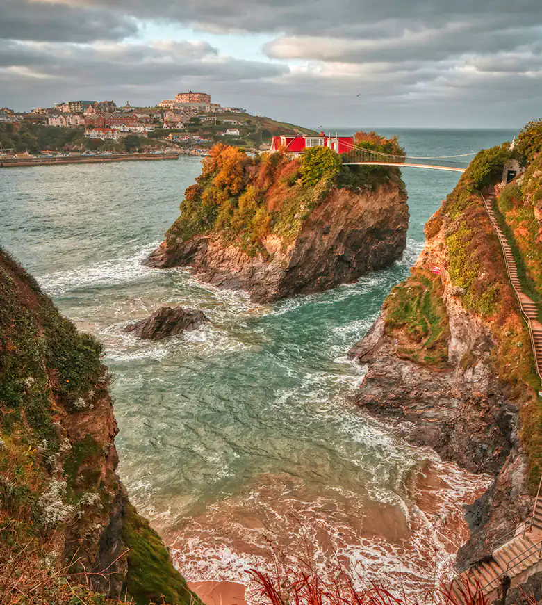 A red house perched on a cliff, overlooking the ocean in Newquay, Cornwall, with waves crashing below.