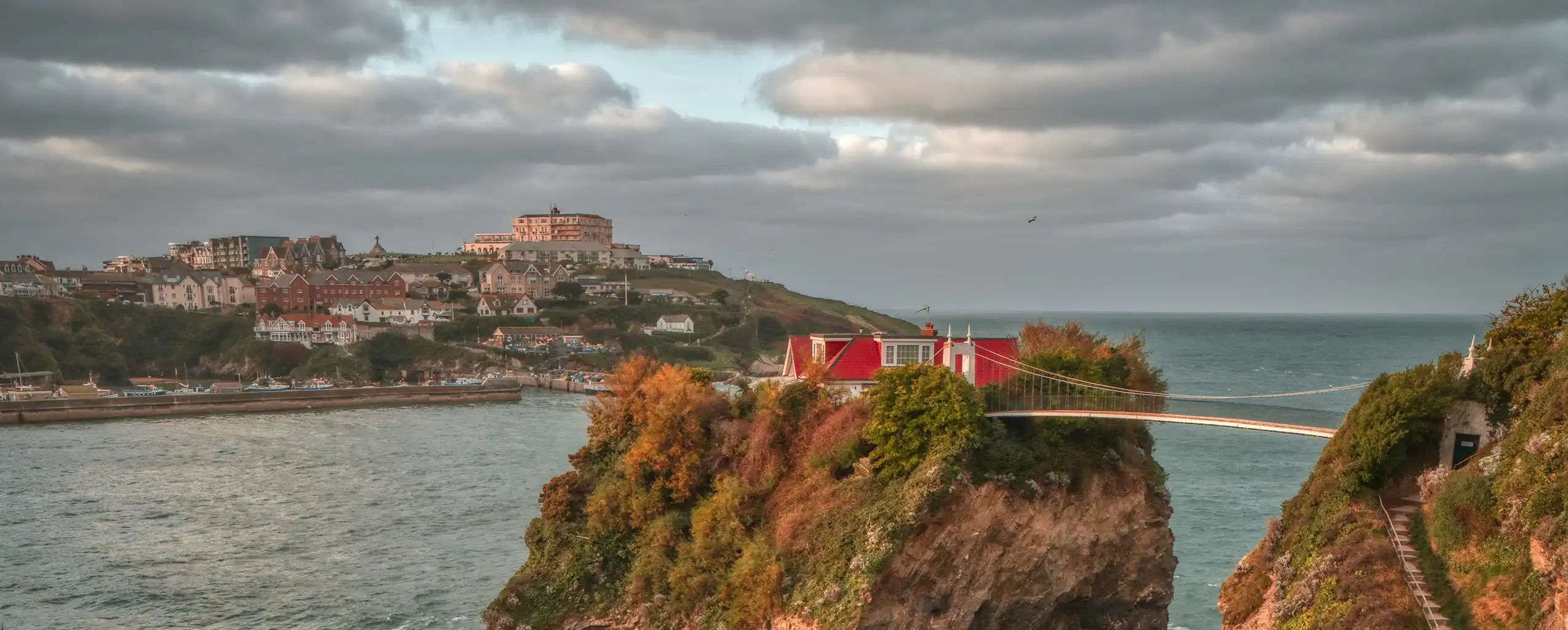 A scenic bridge spans the ocean, with a vibrant red house perched on a rocky cliff in Newquay.