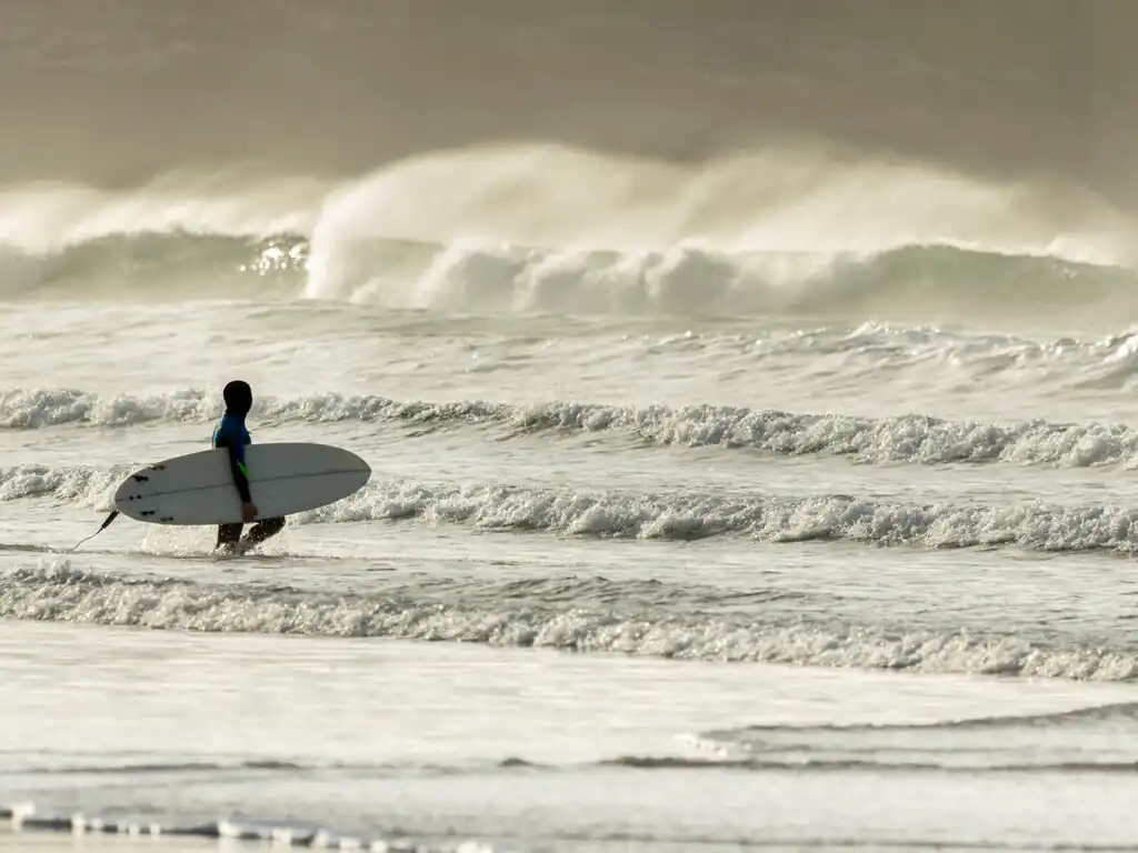 A person walks through the water at Fistral Beach, carrying a surfboard, ready for a weekend of surfing in Cornwall.