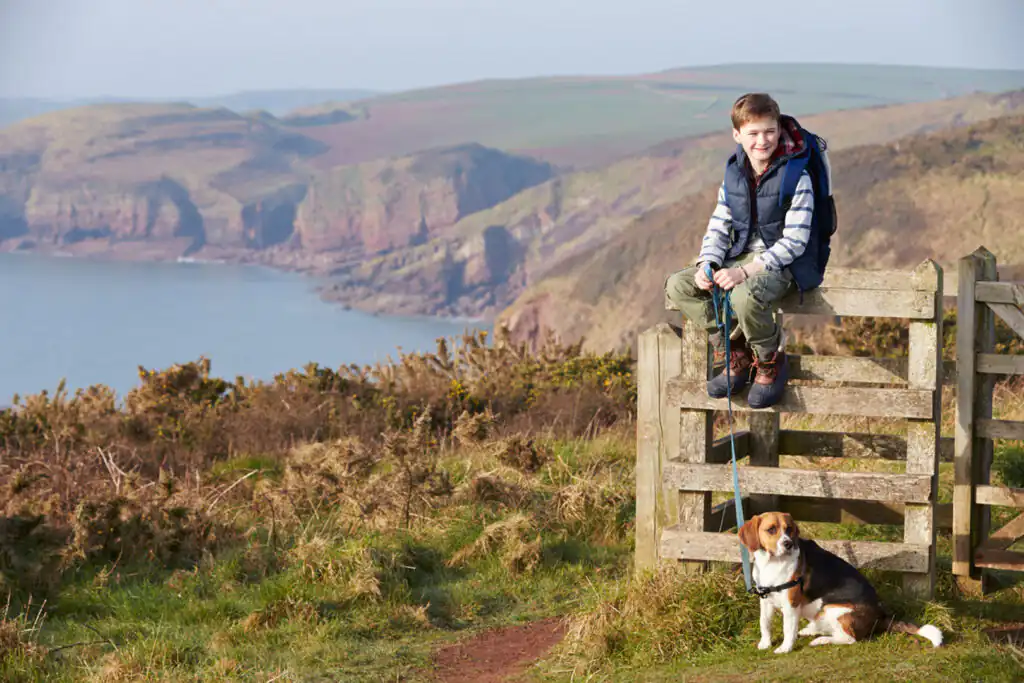 A boy sits on a fence beside a dog, enjoying the view of a cliff path in Cornwall, perfect for dog-friendly adventures.