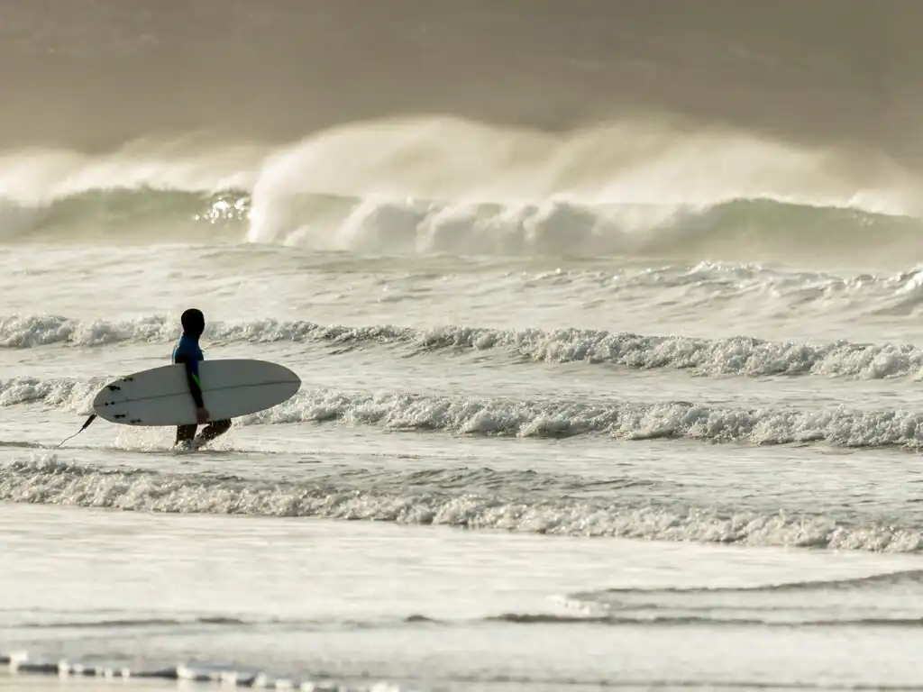 A person walks through the water at Fistral Beach, carrying a surfboard under their arm, ready to catch some waves, a must do activity on a short break in Cornwall.