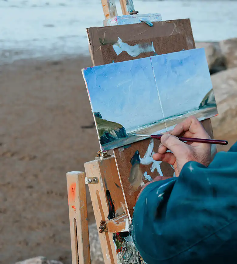 A person painting on Mawgan Porth Beach, with the sandy shore and ocean waves visible in the background.