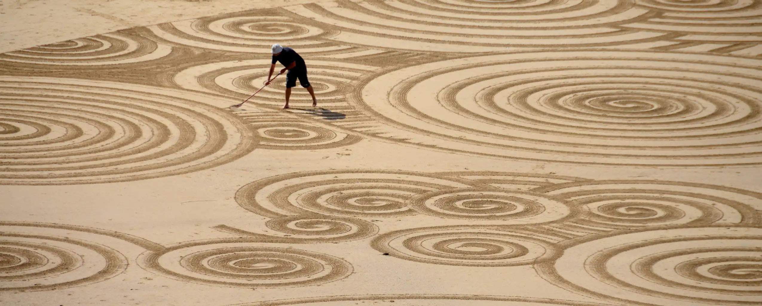 A man stands on Tolcarne Beach in Newquay, proudly displaying a large, intricate sand art creation, a great peice of Newquay art and heritage.