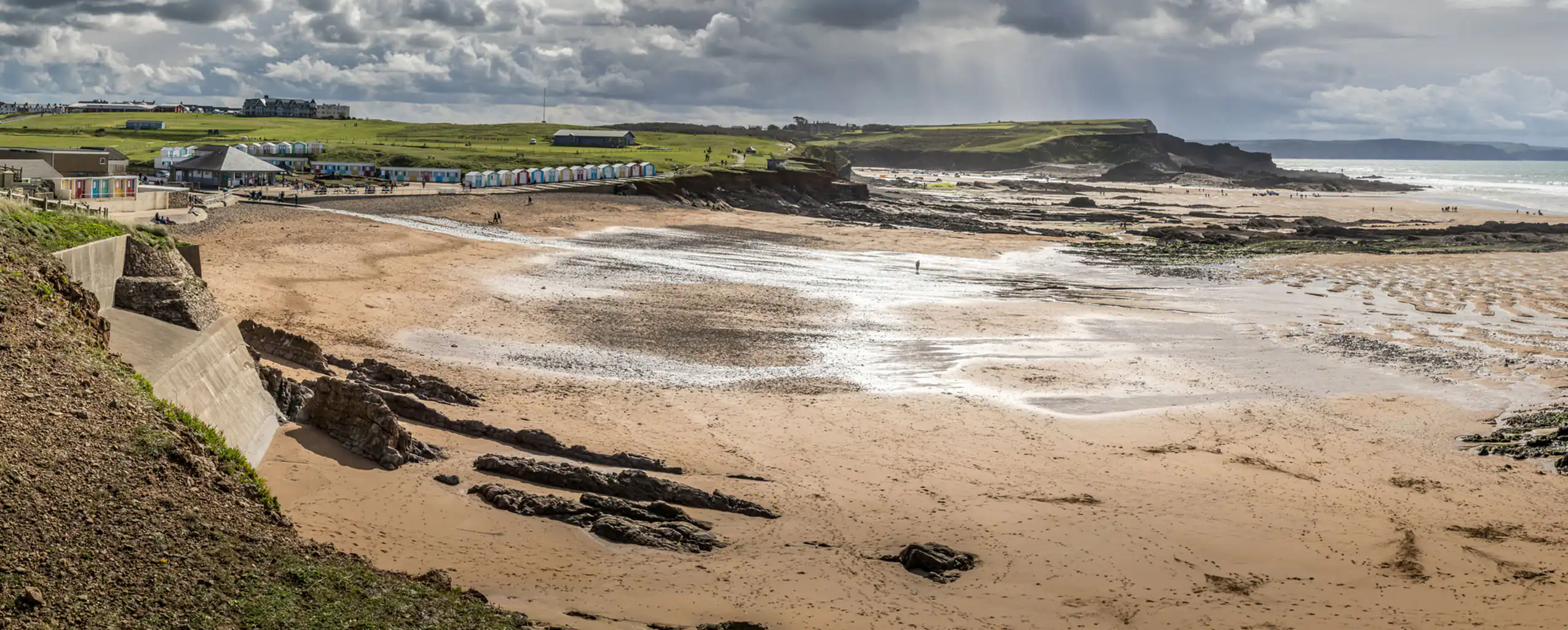 Crooklets Beach in Bude, Cornwall, featuring a rocky shore and a lush grassy area under a clear blue sky. great to visit as a group from a Cornwall cottage sleeping 8.
