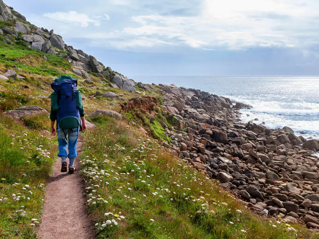 A woman walks along the South West Coastal Path beside the ocean, enjoying the scenic views of the Cornish coastline.