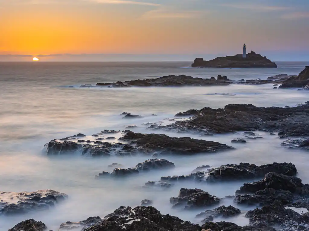 Godrevy Lighthouse at sunset, surrounded by rocky coast and shimmering water in Hayle, a great place to visit. A picturesque coastal scene.