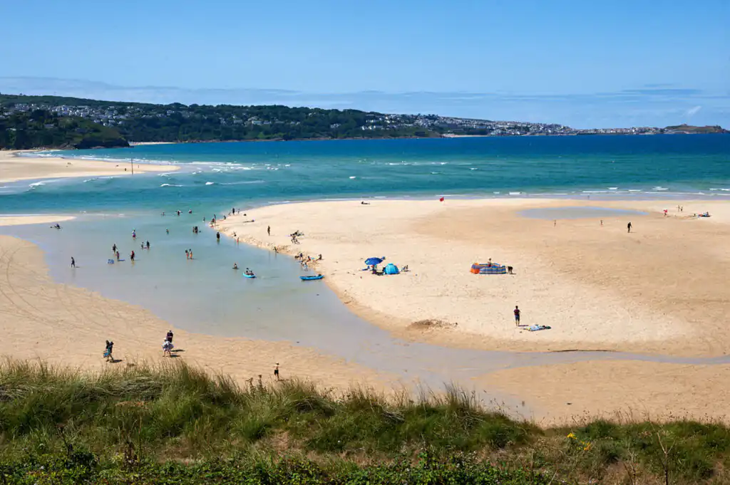 A sandy Mexico Towans beach with people swimming and sunbathing, a shallow stream, grassy dunes, and the ocean in the background, a great family-friendly beach in West Cornwall.