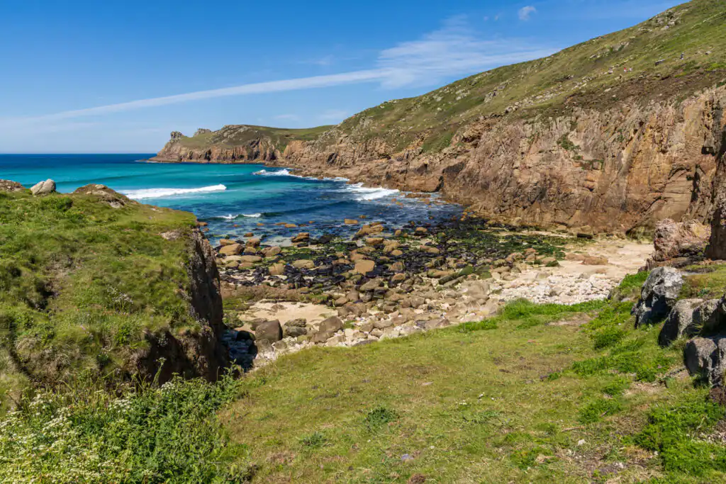 Nanjizal Beach with rocky cliffs, turquoise sea, and grassy foreground under a blue sky, a great hidden beach in Cornwall.
