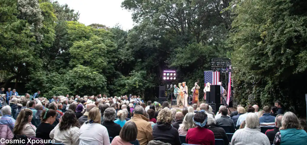 A lively crowd gathers in Penlee Park, Cornwall, enjoying a performance at the open-air theatre, surrounded by greenery.