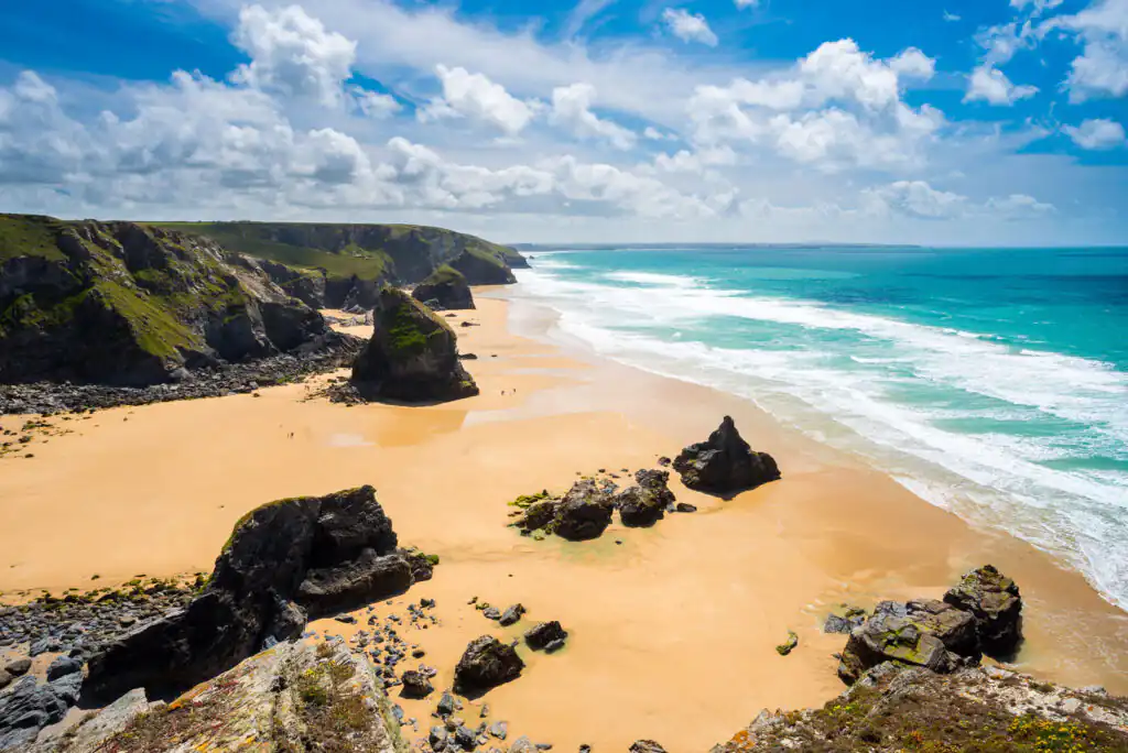 Pentire Steps Beach with sandy shore, rocky formations, and cliffs under a blue sky with clouds, a great, lesser-known hidden beach in Cornwall.