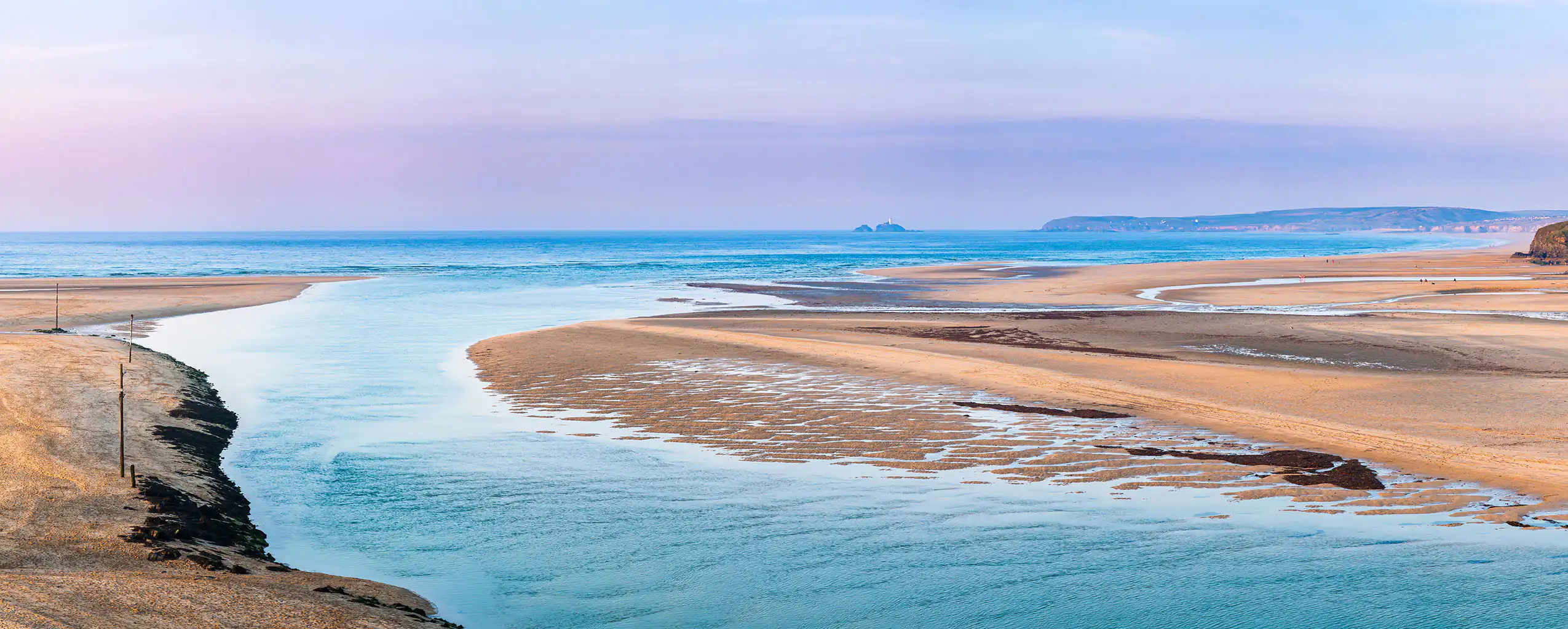 A serene river flows through the sandy expanse of Porthkidney Beach in Lelant, blending nature and tranquillity, a great place to bring some food and drink.