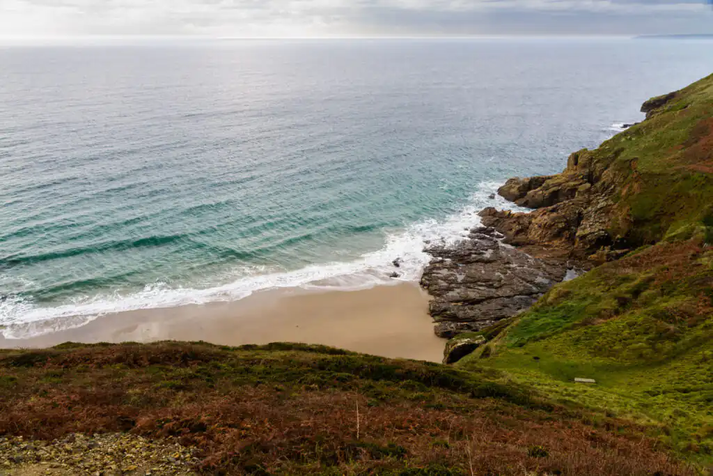 Rinsey Cove in Cornwall with sandy beach, rocky cliffs, and turquoise sea under an overcast sky, a great hidden cove in Cornwall.