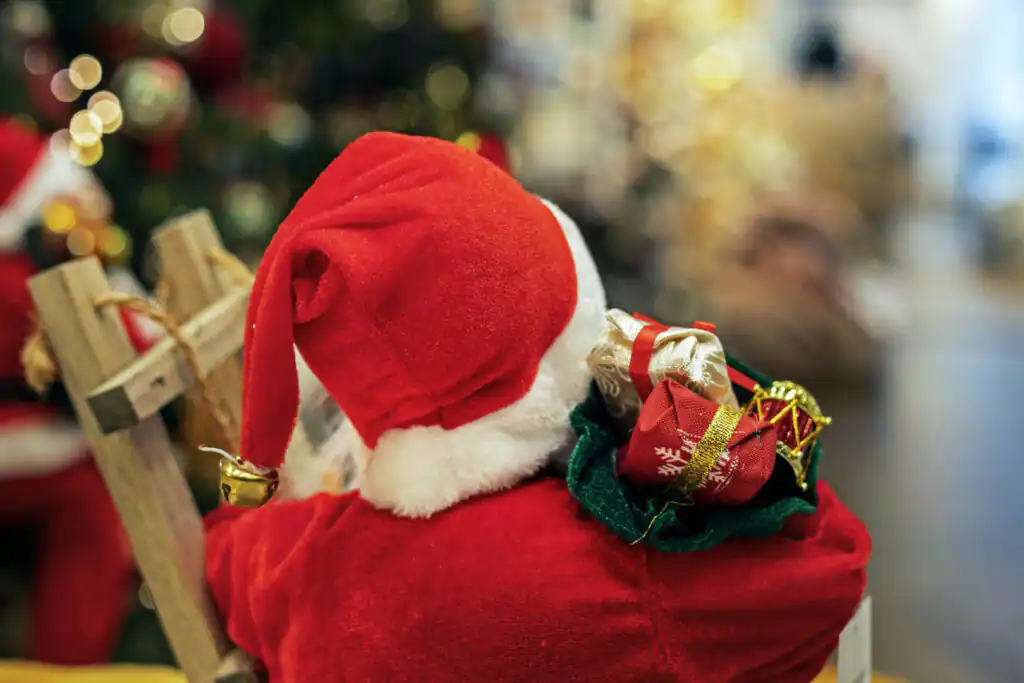 Santa Claus doll holding presents sits on a chair, with a decorated Christmas tree in the background, celebrating Christmas in Cornwall.