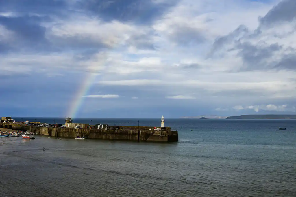 St Ives Harbour with a lighthouse on a stone pier, a rainbow, and boats on the water, a great thing to do in winter in St Ives.
