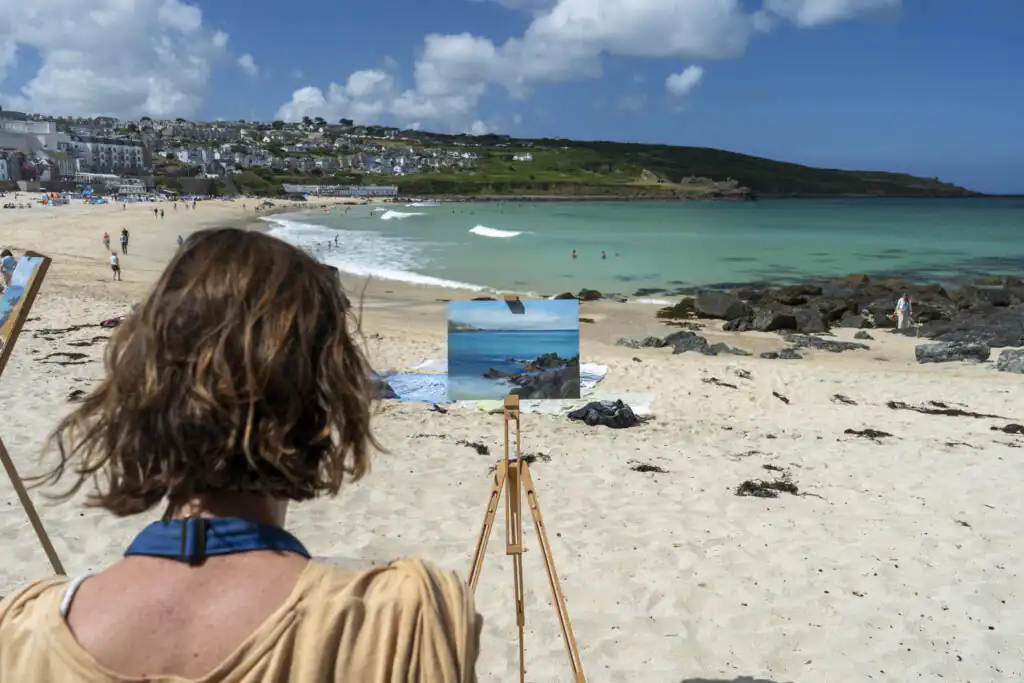 A woman immersed in painting on the sandy shores of Porthmeor Beach, part of the St Ives School of Painting.