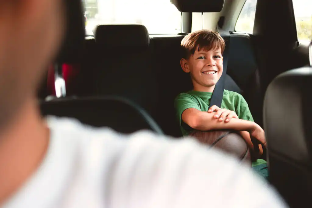 A smiling boy in a green t-shirt, sitting in a car's back seat with a seatbelt on, playing with a ball on his car journey to Cornwall.