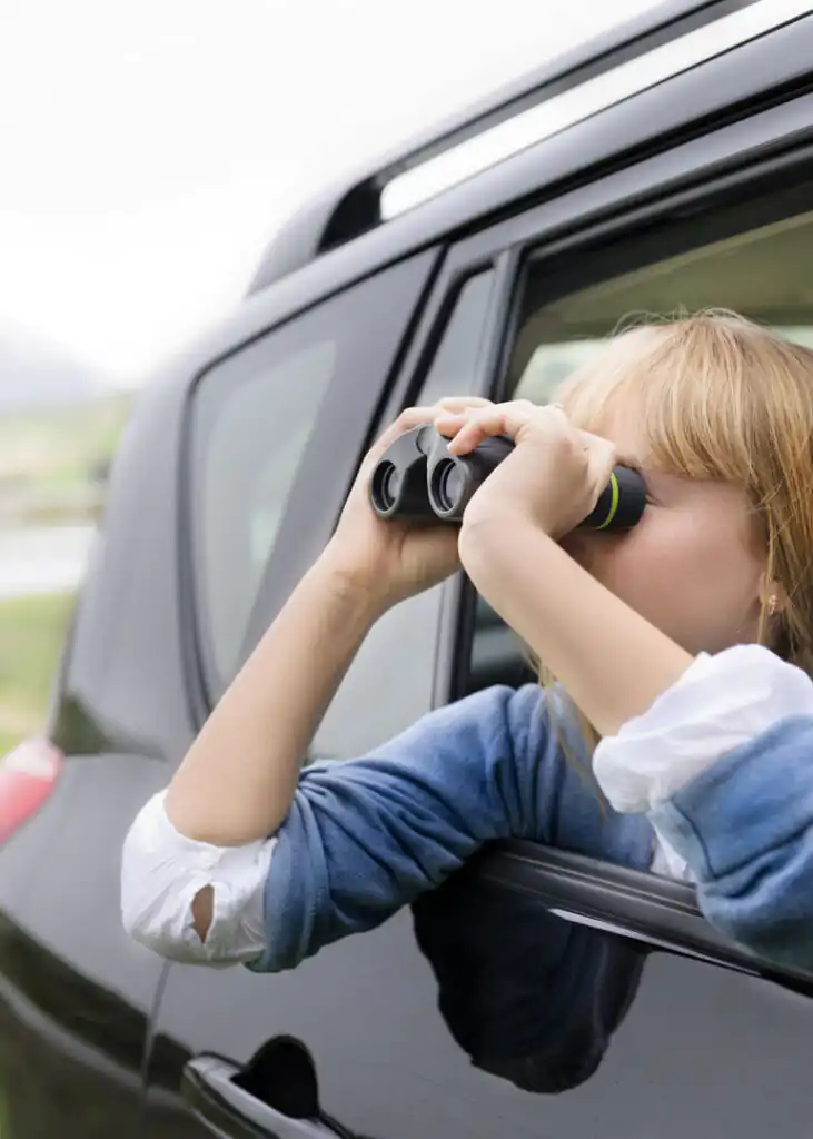 Child looking out of a black car window with binoculars, on their car journey to Cornwall.