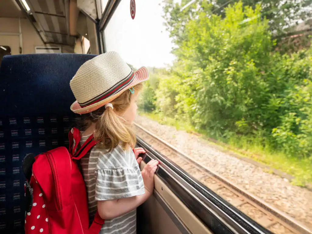 A little girl in a hat and backpack gazes out the train window, enjoying the scenic views of a Cornwall rail journey.