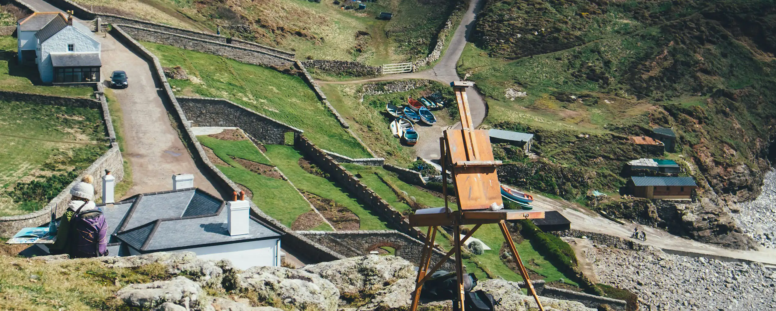 Overlooking a house and beach, an easel stands on a hill capturing the beauty of Cape Cornwall's landscape and Mounts Bay's arts and heritage.