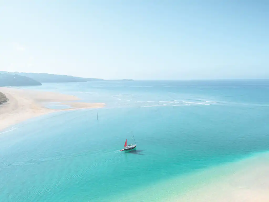 A sailboat with a red sail on the turquoise waters of Hayle Estuary, with a sandy beach and distant coastline, just one location in which Cornish Escapes can help with holiday cottage ownership.