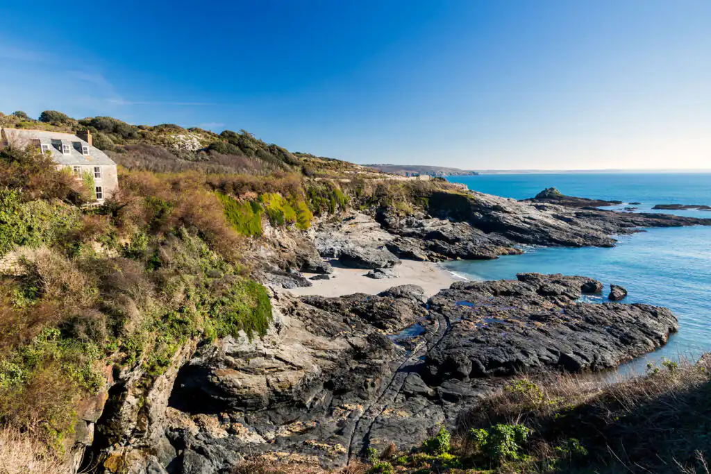Coastal landscape of Prussia Cove, Cornwall, with rocky cliffs, a sandy beach, and a stone building surrounded by greenery, a great hidden cove in Cornwall.