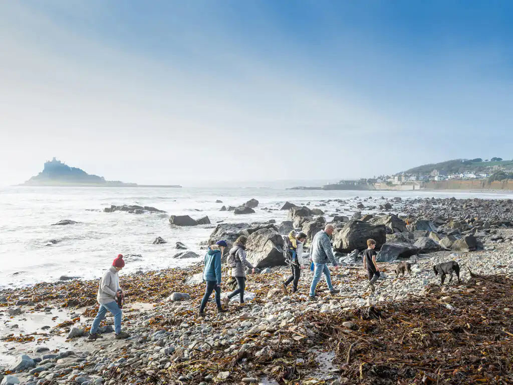 A group of people strolls along Marazion Beach, enjoying the sun and walking a happy dog by the shoreline, a great thing to do in Marazion.