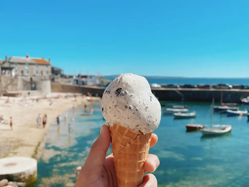 A person enjoys an ice cream cone by the ocean in Mousehole, Cornwall, with waves gently lapping in the background, a great example of Mounts Bay's food and drink.