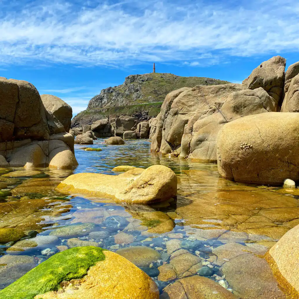 Rocky shoreline with water, a lighthouse in the background, and a rockpool, showcasing Cornwall's coastal beauty and a great place for a walk to learn about Cornwall's geology.