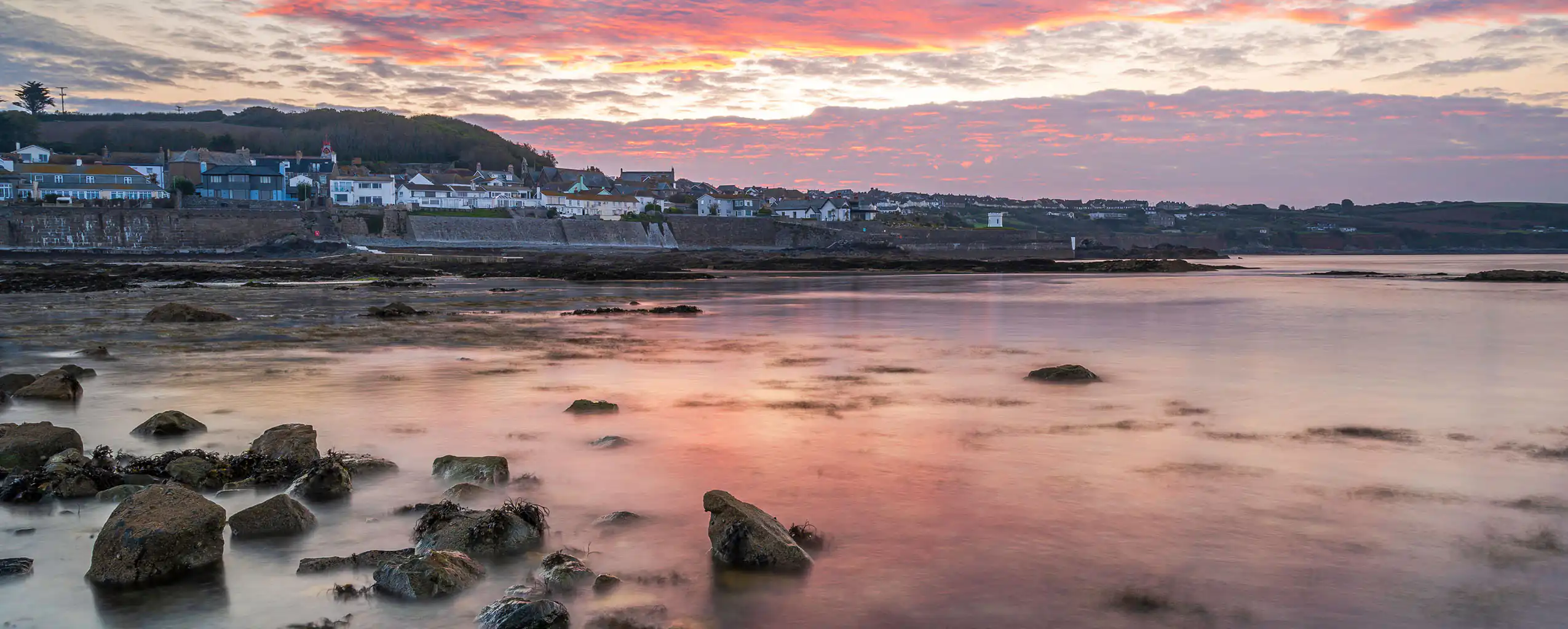 A vibrant sunset casts warm colors over a rocky shoreline, with distant houses silhouetted against the sky in Marazion, a great place for food and drink in Cornwall.