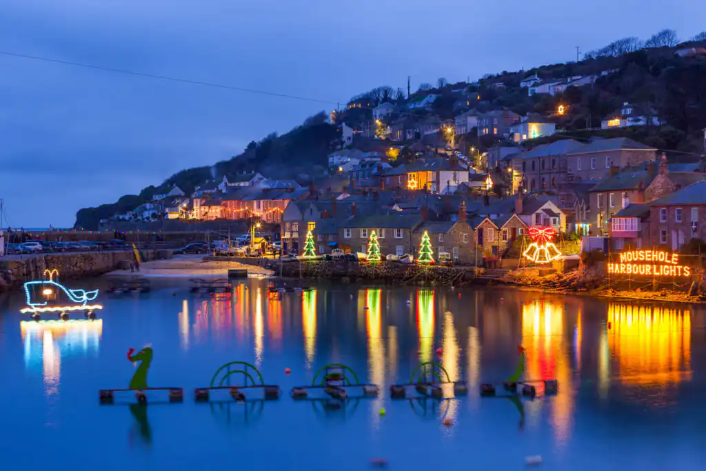 Mousehole harbour illuminated with colourful Christmas lights, featuring boats gently bobbing on the shimmering water, a great place to visit for Christmas in Cornwall.