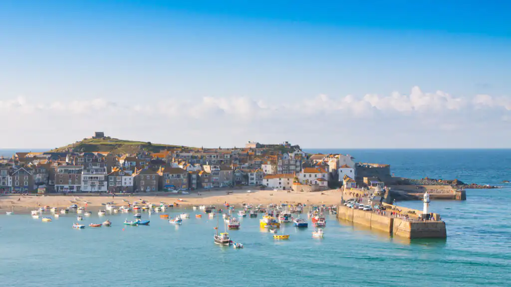 St Ives Harbour with colourful boats, sandy beach, historic buildings, and a lighthouse on a stone pier under a blue sky, a great place for holiday cottage ownership.